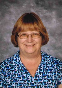 Ms. Mary Horvath is pictured on a gray background. She has short medium-blond hair, wears glasses, and is wearing a blue patterned shirt.